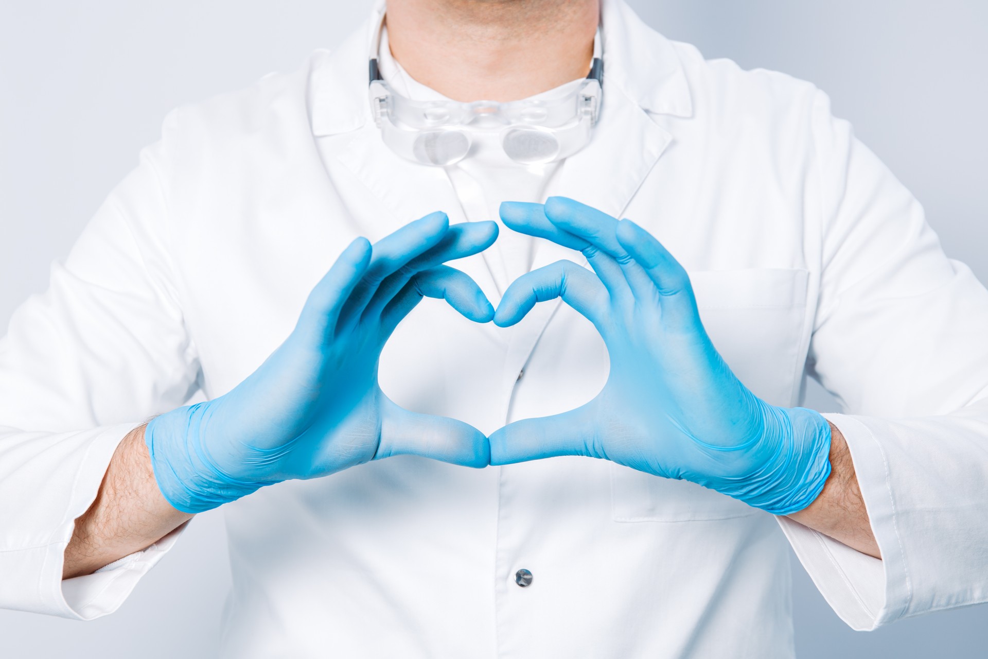 Doctor hands in gloves in the heart shape against the background of his body and medical gown. closeup. Male hand in blue medical gloves show heart sign.